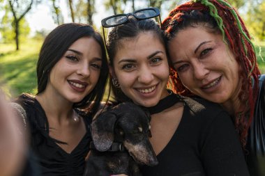 Üç gülümseyen kadın ve köpek dışarıda eğleniyor, grup selfie anı yakalıyorlar.