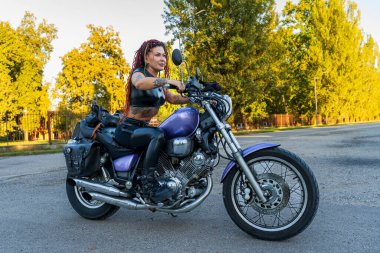 Yolda motosiklet süren rastalı ve dövmeli bir kadın.