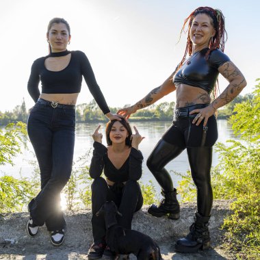 Üç alternatif kadın ve küçük bir köpek nehir kenarında poz veriyorlar.