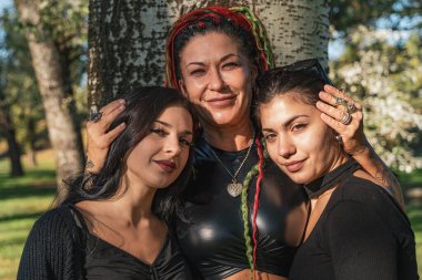 Eşsiz tarzları olan üç kadın açık havada kucaklaşıyor, aile bağlarını gösteriyor.