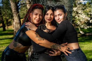 Kadın arkadaşlar ağacın yanında evcil köpekle poz veriyorlar, dostluk ve hayvanlarla arkadaşlık gösteriyorlar.