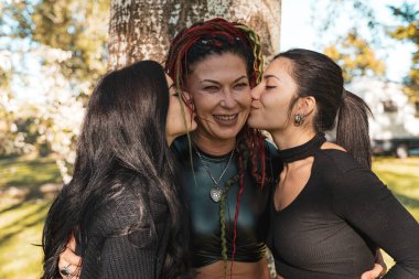 Mutlu annelerini iki yanağından öpen iki genç kadın bir ağacın önünde.