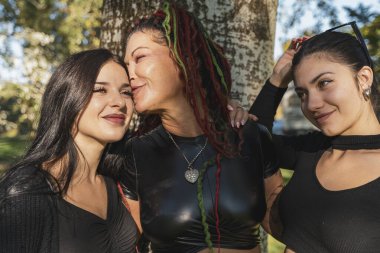 Anne dışarıda kızlarını kucaklıyor ve öpüyor, sevgisini ve mutluluğunu paylaşıyor.