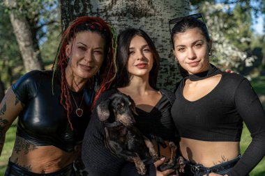 Rastalı ve dövmeli kadınlar parkta dachshund ile poz veriyorlar.