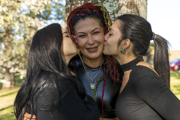 Mutlu annelerini iki yanağından öpen iki genç kadın dışarıda sevgi ve şefkat gösteriyorlar.