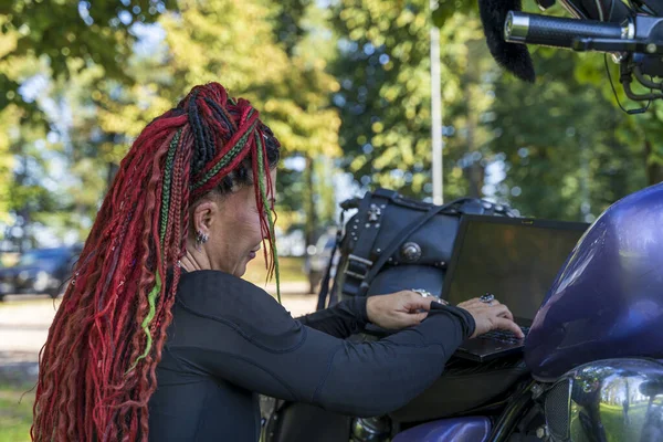 Motosikletinin üzerinde otururken dizüstü bilgisayarda yazan renkli rastalı bir kadın bisikletçi.
