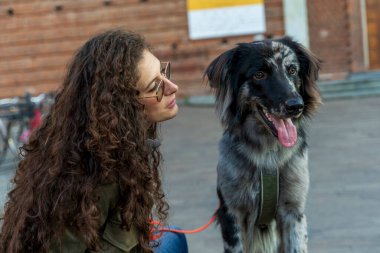 Kıvırcık saçlı bir kadın şehirde köpeğine bakıyor, yakın bağlarını gösteriyor.