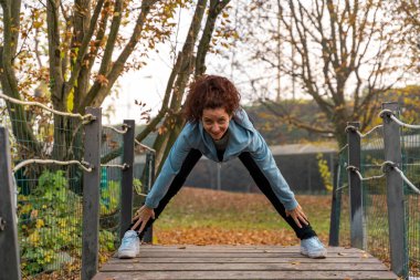 Bir sonbahar parkındaki tahta bir köprüde bacaklarını esneten bir kadın.