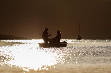 A boat on the water at sunset with two anglers against the light clipart