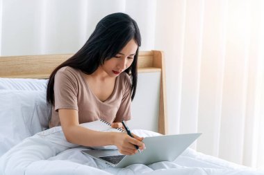 Teknoloji konseptiyle evde yaşam tarzı çalışması. İnternette çalışan serbest çalışan genç bir kadın sabah evde yastıklı beyaz yatakta otururken kitaba bir şeyler yazıyor.. 