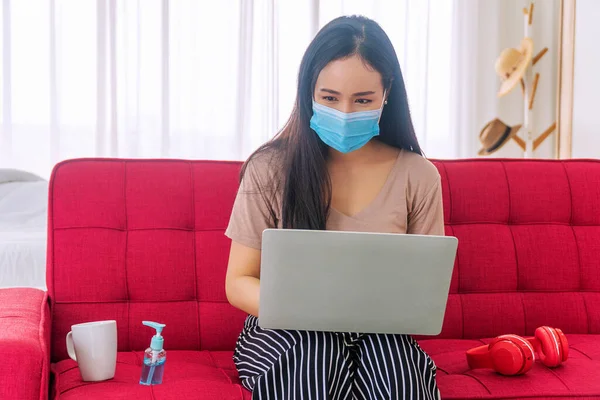 Healthy disease lifestyle concept. Young woman wearing surgical protective mask over her face using laptop computer working from home while sitting on red sofa with sanitizer alcohol and headphone.