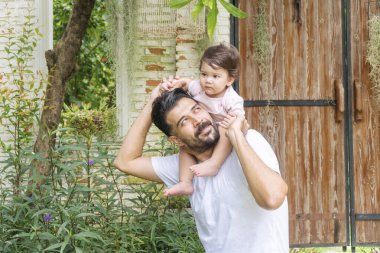 Mutlu Babalar Günü konsepti. Neşeli baba evde dinlenirken küçük kızını boynuna alıp sırtında taşıyor. Bahçede babacığının omzunda küçük sevimli melez bir kız. . 