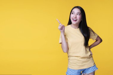 Neşeli genç kadın, sarı arka planda izole bir şekilde dikilirken ürünü veya boş kopya alanını işaret ederek parmak kullanıyor.