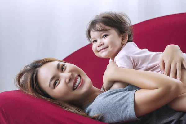 遊び心のある若い母親と混合レースの娘は リビングルームで赤いソファの上に一緒に横になりながら楽しみを持っています — ストック写真