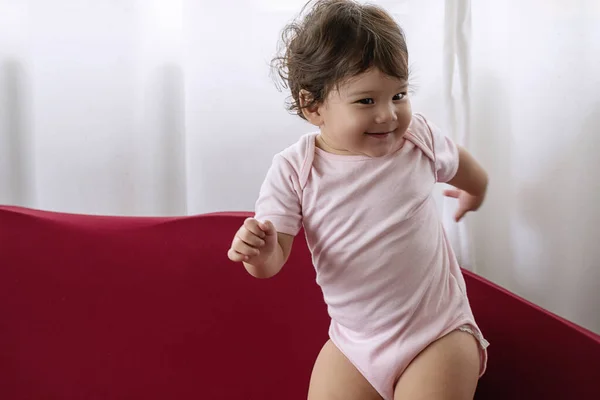 Little cute baby girl play on red sofa at home. Enjoy one year old child mixed race smiling while standing on the couch. Adorable toddler wear pink bodysuits dancing on sofa cushion in living room.