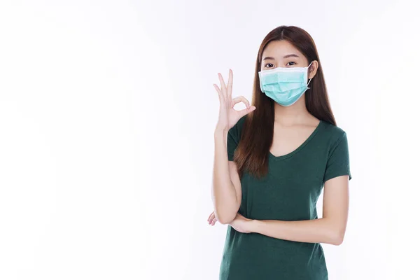Güvenli tıbbi maske takan genç bir kadın havadaki bakteri, virüs hastalığı ve hava kirliliği sorununu burnuna solumaktan korunurken el işaretini beyaz arka planda iyi yönde sağlıklı hale getiriyor..