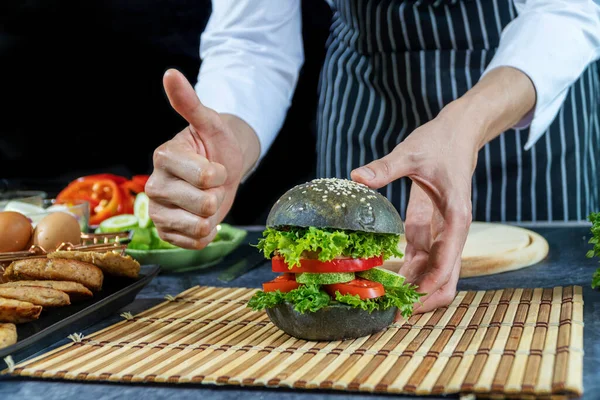 Ev yapımı ekmek burger konsepti. Şefin eli, siyah arka planda sebzeli hamburgerle baş parmağınızı kaldırın.. 