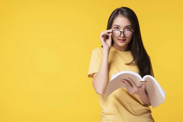 Yaşam tarzı hobisi ya da bilgi konsepti. Güzel genç bir kadın, elinde kitap okurken komik gözlüklerini çıkararak izole edilmiş sarı arka planda aldatıcı görünüyor.. 