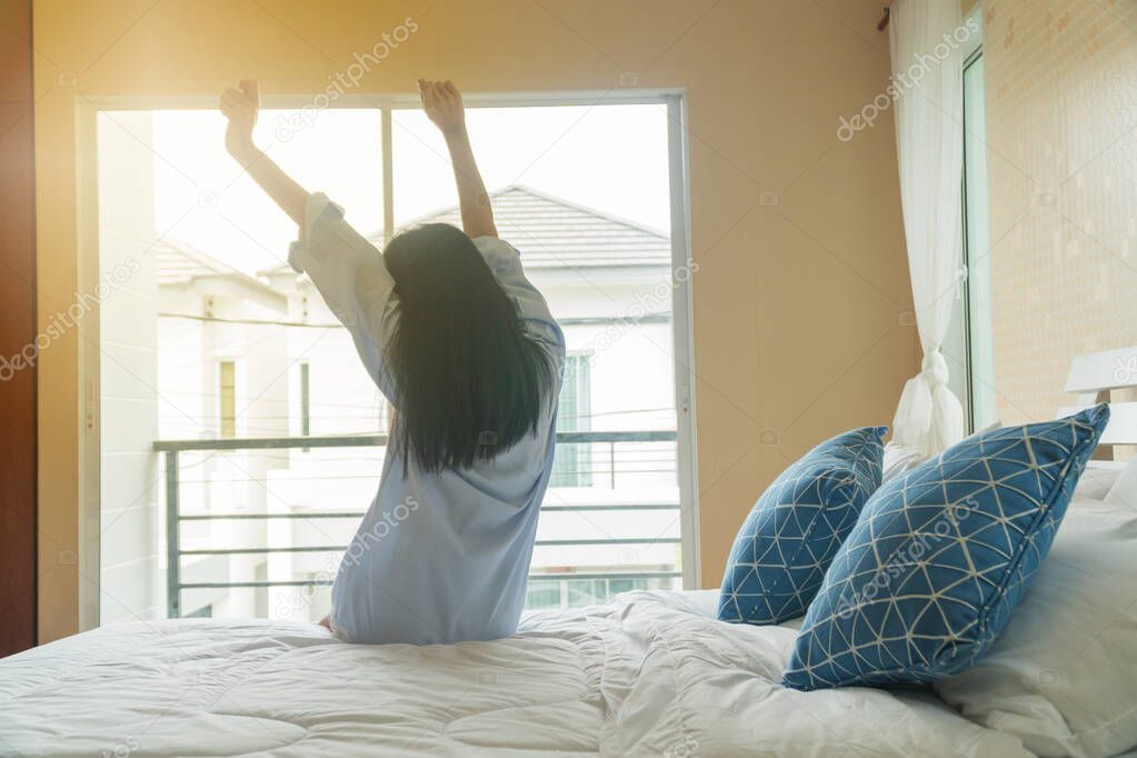 Rear view of beautiful Asian young girl sitting stretched in bed. Facing the balcony in the morning.