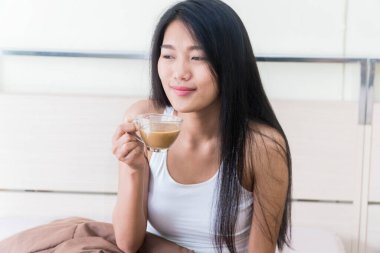 Güzel, genç Asyalı bir kadın elinde bir fincan kahveyle yatakta oturuyor..