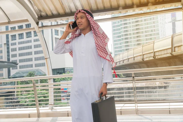 讲手机的穆斯林商人 年轻的阿拉伯商人在一个现代化的城市里拿着公文包 用智能手机的生活方式来交谈 — 图库照片