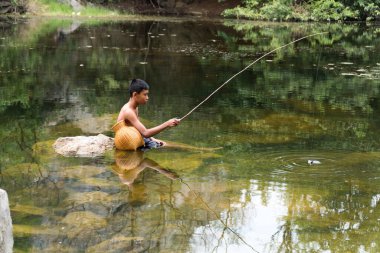 2017 Ekim Ratchaburi, Taylandlı çocuk gölette olta atıyor. Göletin ortasında mavi pantolon giyen çocuk kayanın üzerinde oturuyor, arka planda oltanın ucunda balık yakalıyor..