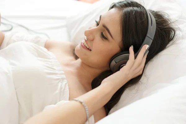 美しい笑みを浮かべて若い女性の白いベッドのヘッドフォンを着て音楽を聞くに横たわっている ライフスタイルコンセプト — ストック写真