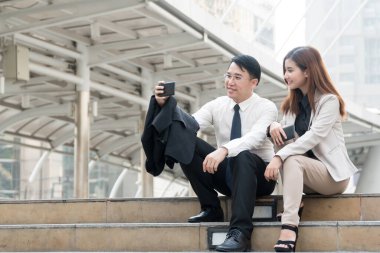 Merdivenlerde oturan iki işadamı modern şehirde ellerinde akıllı telefonlarıyla selfie çekiyorlar. Merdivende otururken mutlu mesut selfie çekeriz.