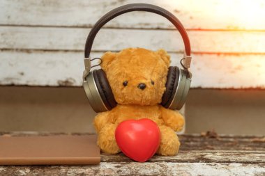 Kırmızı kalpli kulaklık takan kahverengi oyuncak ayı bahçede bir sandalyede oturuyor..