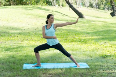 Parklarda yoga yapan orta yaşlı, beyaz bir kadın. Parktaki çimlerin üzerinde yoga minderi üzerinde duran kadın dışarıda egzersiz yapıyor. Sağlıklı yaşam tarzı kavramı.