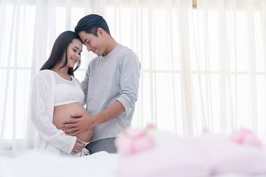 Koca beyaz pencerenin yanında karısına sevgiyle sarılıyor. Mutlu bir çift genç adam hamile güzel karısına sarılıyor, gülümseyerek ve birbirlerinin gözlerinin içine bakarak. Bebek sahibi olma kavramı.