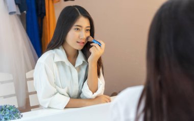 Aynanın karşısında oturan mutlu genç kadın odada makyaj yapıyor. Çekici Asyalı kadın fırça tutuyor ve aynanın önünde otururken makyaj yapıyor..