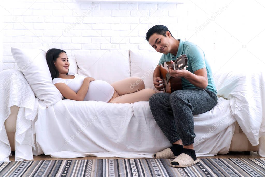 Young husband play the guitar for his smiling pregnant wife listened happily on white couch in living room. Young man playing acoustic guitar for his pregnancy wife relax at their house.