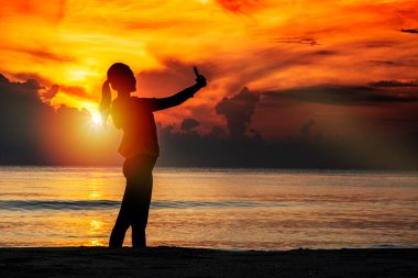 Akıllı telefon kullanıp sosyal medyada selfie çeken genç bir kadının silueti. Sabah güneş doğarken plajda rahatlamak. Tatil tatili doğa deniz konseptiyle yapılır..