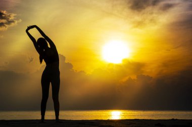 Yaşam tarzı rahatlama egzersizi konsepti. Siluet genç kadın sabah güzel bir gündoğumuyla sahilde dururken ince vücudunu esnetiyor..