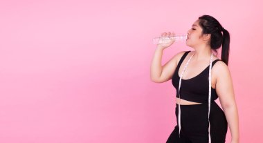 Aşırı kilo kaybı konsepti. Şişman, şişman, genç bir kadın. Elinde mezuralarla idmandan sonra elindeki şişeden taze su içerken izole edilmiş pembe arka planda duruyor..