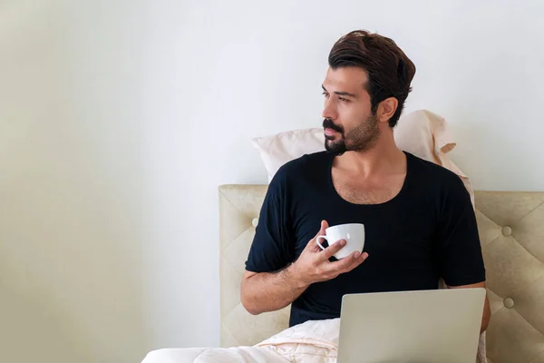 Yaşam tarzı karantina konsepti. Yakışıklı serbest çalışan genç adam, yatakta otururken bilgisayarını kullanarak siyah tişört giyiyor. Sabahları elinde kahve ya da çay tutuyor ve içiyor.. 