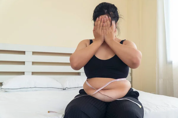 Obezite sağlıksız bir kavram. Kilolu, Asyalı bir kadın, beyaz yatakta otururken el ele tutuşup evinde yüzünü kapatıyor. Yatak odasındaki ekstra ağırlıktan muzdarip bir kadın mezurası..