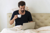 Karanténní koncept životního stylu. Fešák na volné noze mladý muž nosí černé tričko pomocí notebooku pracuje on-line, zatímco sedí na posteli. Ráno drží a pije šálek kávy nebo čaje. 