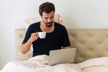 Yaşam tarzı karantina konsepti. Yakışıklı serbest çalışan genç adam, yatakta otururken bilgisayarını kullanarak siyah tişört giyiyor. Sabahları elinde kahve ya da çay tutuyor ve içiyor.. 