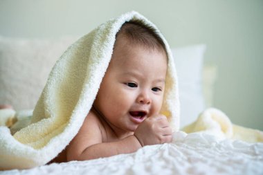 Neşeli şirin bebek beyaz battaniyenin altında kameraya bakıyor. Masum bebek, beyaz yatakta kafasında havluyla sürünüyor..