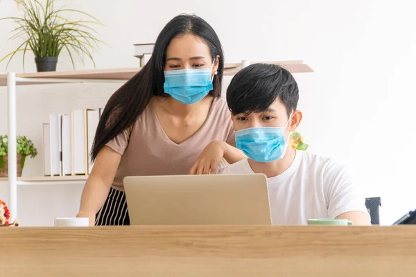 New normal work from home, health care, covid outbreak concept. Freelance young couple wear wearing face mask use laptop working or checking newscast or searching information together in room at home.