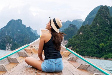 Maceracı hayat tarzı tatilcisi rahatlama konsepti. Gezgin genç kadın şapka takıyor uzun kuyruklu teknede oturuyor dağlarla ve gökyüzen gölde yüzerken Surthani 'de tatil turisti.