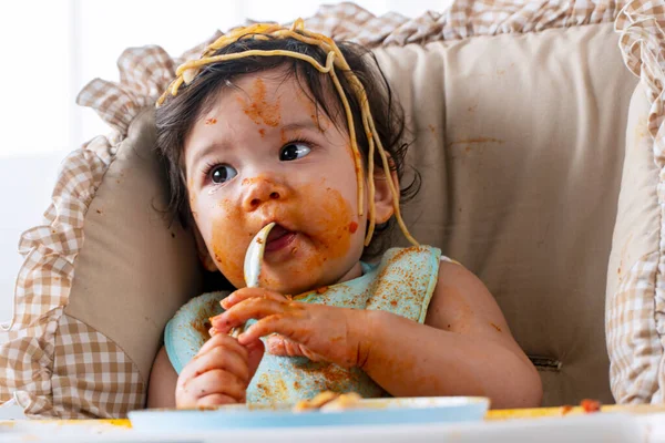 12 aylık sevimli, komik bir kız kaşıkla spagetti yerken evinde güçlü bir sandalyede oturuyor. Yüzünde domates sosu olan küçük bir çocuk ailesine bakıyor. Kendini besleme konsepti