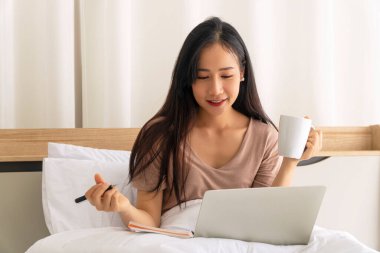 Serbest çalışan genç bir kadın, sabahları elinde yastıkla beyaz bir yatakta otururken elinde bir fincan kahveyle dizüstü bilgisayarda çalışıyor. Teknoloji konseptiyle evde yaşam tarzı çalışmaları.