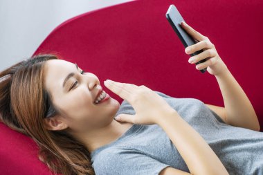 Yaşam tarzı teknoloji konsepti. Oturma odasındaki kırmızı kanepede uzanırken akıllı telefon tutan neşeli genç kadın. Sosyal medyada cep telefonuyla mesaj bağlayan sıradan bir kız..