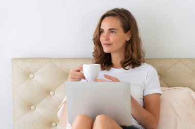 Yaşam tarzı karantina konsepti. Yakışıklı, serbest çalışan genç bir kadın, beyaz tişört giyiyor. İnternette çalışırken yatakta oturuyor. Sabahları elinde kahve ya da çay tutuyor ve içiyor..