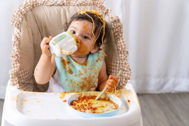 Evdeki güçlü sandalyede otururken kaşıkla spagetti yiyen ve şişeden su içen sevimli küçük komik kız. Yüzünde domates sosu olan küçük bir çocuk. Kendini besleme konsepti.