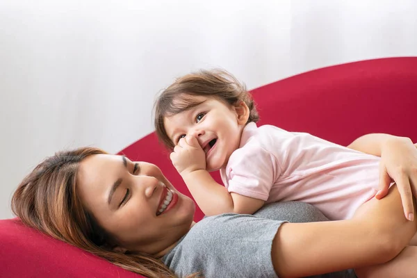 母の日のコンセプト 若い母親のキス混合レースの娘はリビングルームで赤いソファの上に一緒に横になりながら楽しみを持っています — ストック写真