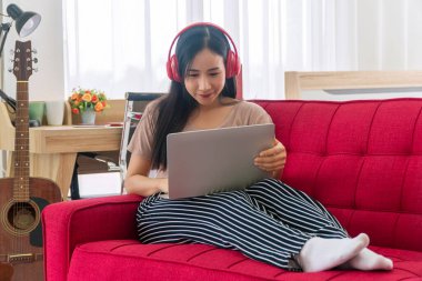 Yaşam tarzı teknolojisi ya da ev konseptindeki yeni normal çalışma. Sıradan bir genç kadın internette çalışırken kulaklık takıyor ya da evdeki kırmızı koltukta otururken haber izliyor..